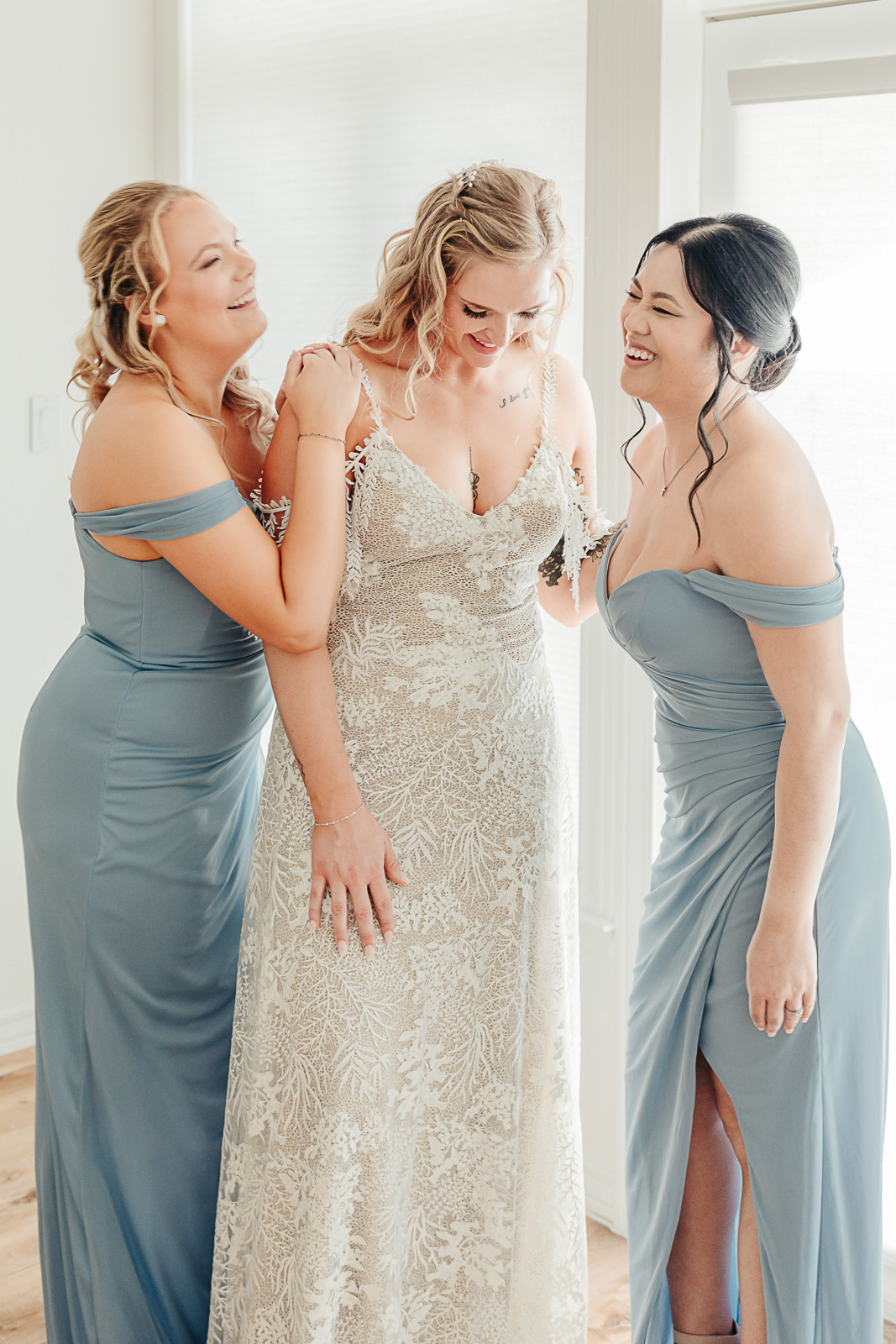 Bride laughs with two bridesmaids in dusty blue off-shoulder gowns while getting ready — Austin Hill Country wedding