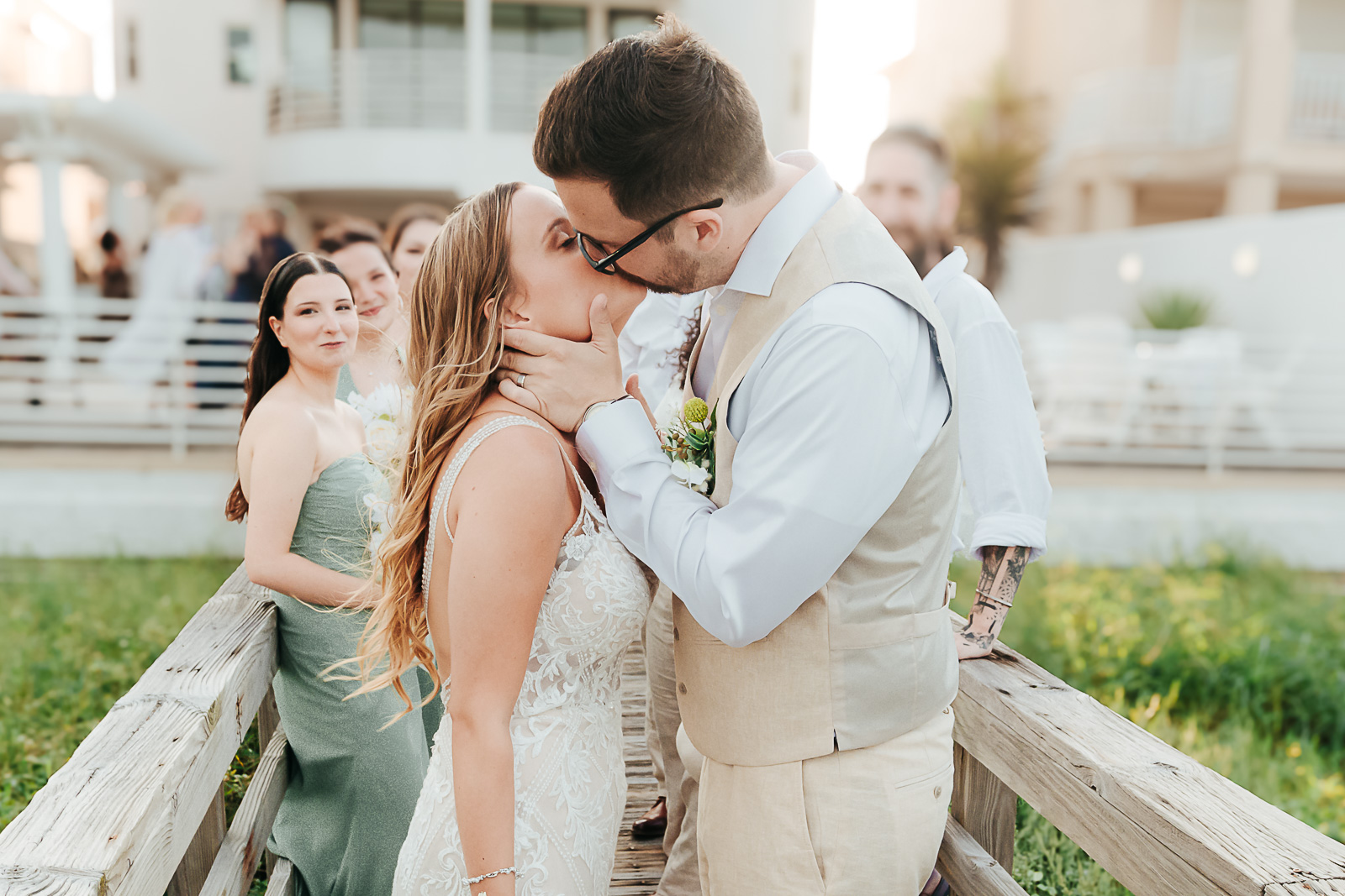 Groom kisses bride on a weathered boardwalk as bridesmaids in sage gowns celebrate — Hill Country wedding ceremony